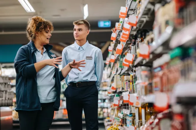 Eine erfahrene Supermarktmitarbeiterin erklärt einem jungen männlichen Auszubildenden die Warenplatzierung in einem Supermarktgang. Beide tragen Dienstkleidung und befinden sich vor einem Regal mit Lebensmitteln.