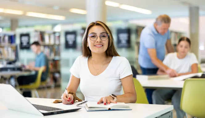 Eine junge Frau mit Brille und weißem T-Shirt sitzt lächelnd in einer Bibliothek und macht Notizen. Vor ihr liegen ein aufgeschlagenes Notizbuch und ein Laptop.