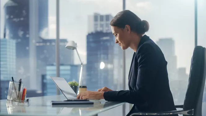 Frau mit schwarzen Haaren in Business Outfit lächelt in ihren Laptop vor einer Fensterfront