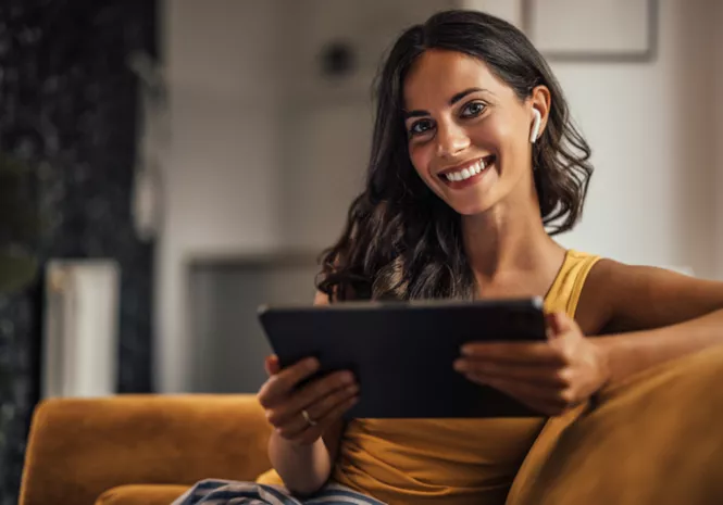 Eine lächelnde Frau mit langen dunklen Haaren und Ohrhörern sitzt entspannt auf einem Sofa und hält ein Tablet in den Händen, während sie in die Kamera schaut.