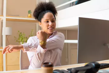 Frau mit schwarzen, lockigen Haaren und pinkem Oberteil dehnt sich am Schreibtisch