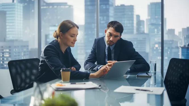 Frau mit schwarzen Haaren in Business Outfit sitzt mit einem Kollegen an einem Schreibtisch vor einem Laptop und bespricht Dinge
