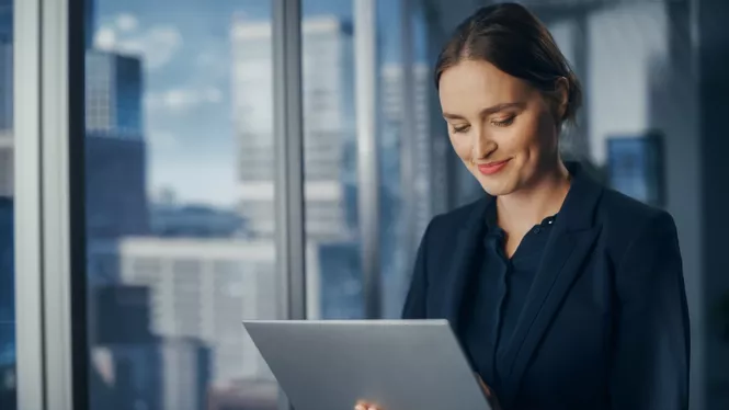Frau mit schwarzen Haaren in Business Outfit lächelt in ihren Laptop vor einer Fensterfront