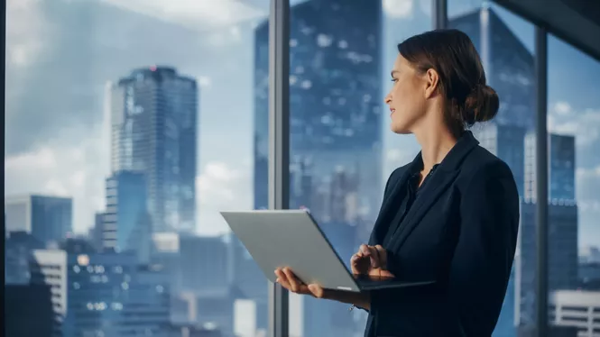 Frau mit schwarzen Haaren in Business Outfit mit Laptop in der Hand schaut aus dem Fenster