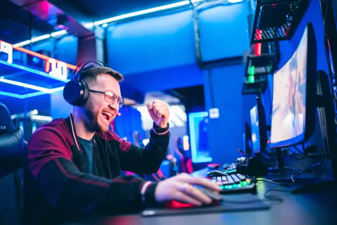 Ein junger Mann mit Brille und Kopfhörern sitzt an einem Computer in einer neonbeleuchteten Gaming-Lounge, lächelt und ballt die Faust vor Freude.