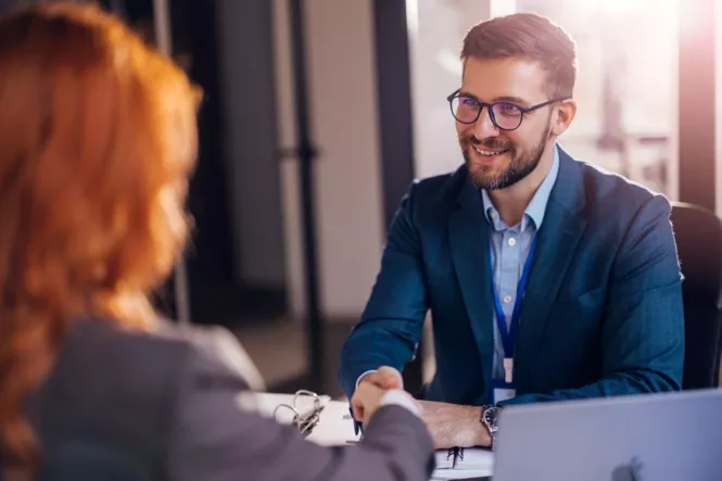 Ein freundlicher Geschäftsmann mit Brille und Bart, gekleidet in einem blauen Anzug, lächelt und schüttelt die Hand einer anderen Person. Die Szene findet in einem modernen Büro bei natürlichem Licht statt.