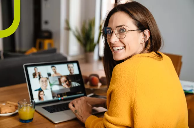 Junge Frau mit Brille lächelt zur Kamera, während sie an einem Laptop mit einer Online-Konferenz sitzt. Neben ihr ein Glas Saft und Kekse.