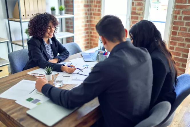 Eine lächelnde Beraterin sitzt mit zwei Kunden an einem Tisch in einem modernen Büro. Vor ihnen liegen Dokumente und ein Laptop, während sie gemeinsam Finanzunterlagen besprechen.