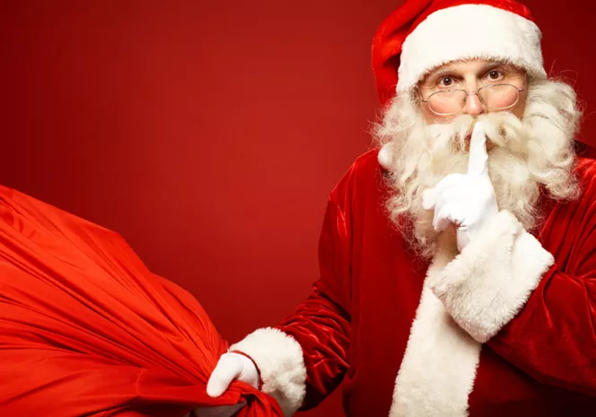 Weihnachtsmann in rotem Kostüm mit weißem Bart hält einen großen roten Geschenkesack und legt einen Finger an die Lippen, vor einem roten Hintergrund.