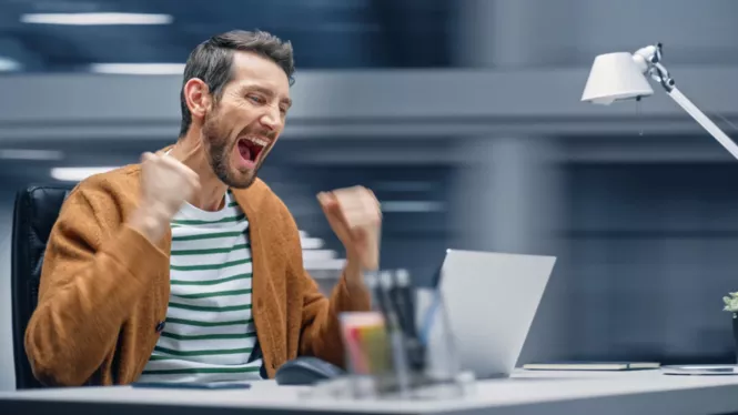 Ein Mann sitzt an einem Schreibtisch vor einem Laptop, hebt die Fäuste triumphierend in die Luft und lächelt vor Freude. Die Szene spielt in einem modernen Büro mit einer Lampe und Bürogegenständen im Hintergrund.