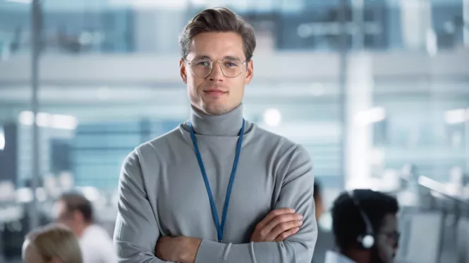 Ein junger Mann im Büro, der ein graues Oberteil trägt, eine Brille und ein Umhängeband um den Hals. Er steht selbstbewusst mit verschränkten Armen vor einer verschwommenen Büroumgebung.