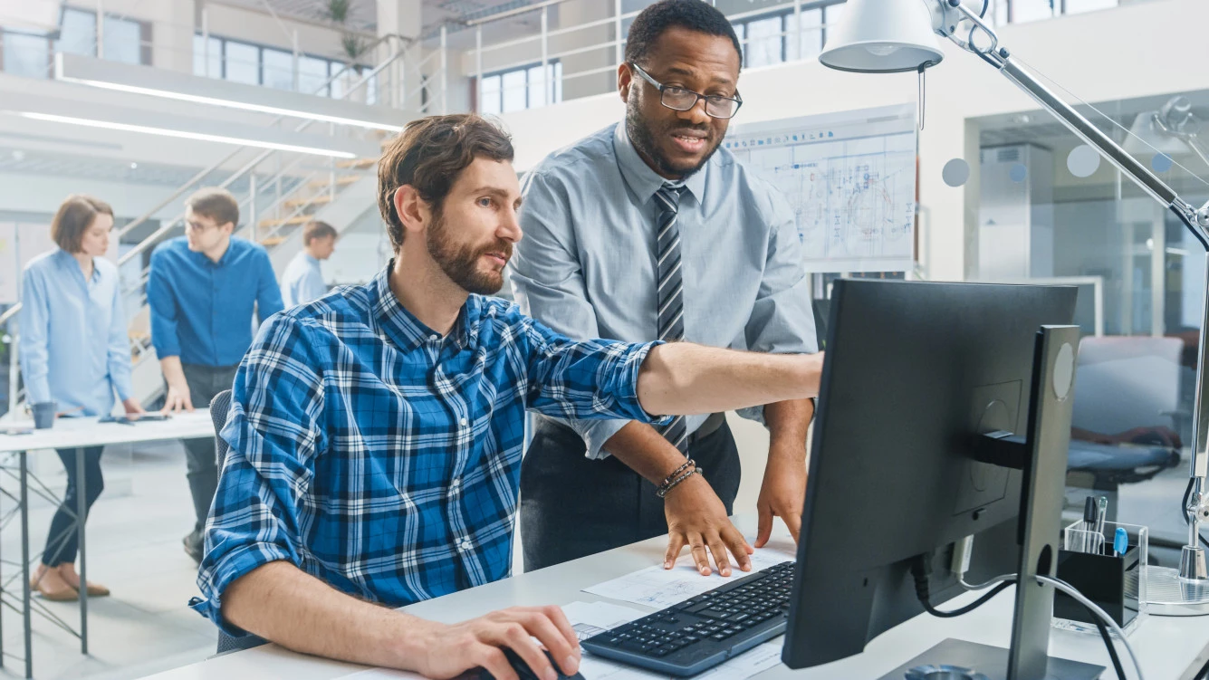 Ein sitzender Ingenieur in blauem, karrierten Hemd zeigt einem Kollegen in grauem Hemd etwas an einem Computer