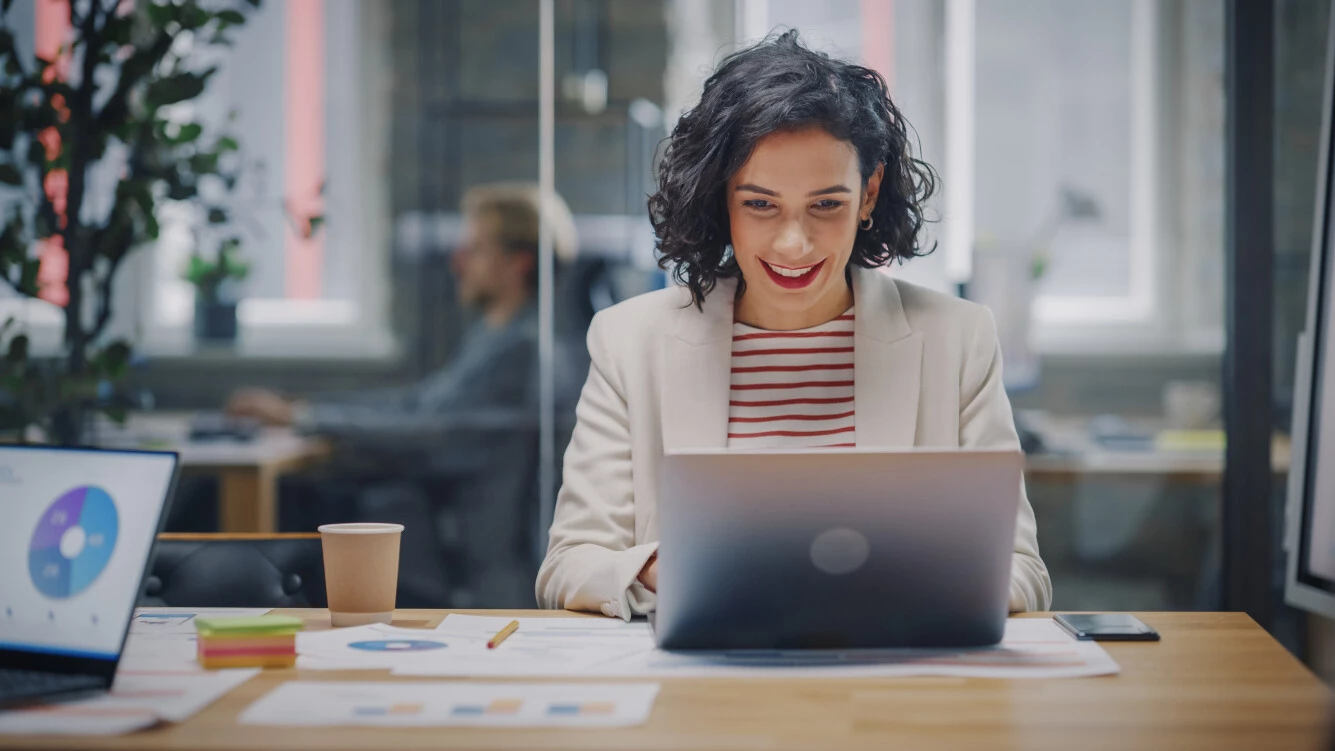 E-Commerce - Frau mit schwarzen Haaren sitzt in modernem Büro an einem Laptop. 